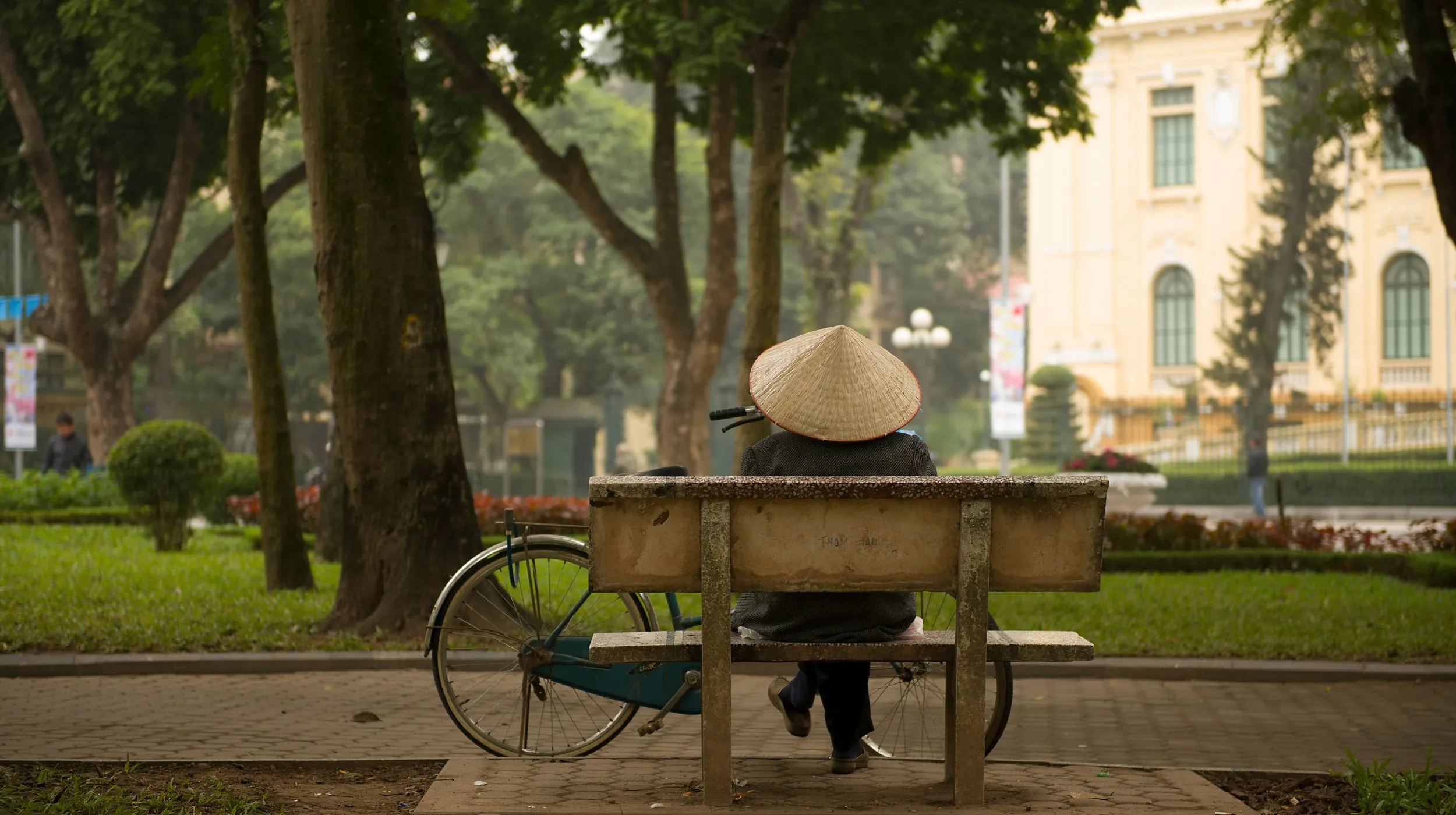 Personne portant un chapeau conique vietnamien assise sur un banc dans un parc de Hanoi, avec un vélo à côté et des bâtiments coloniaux en arrière-plan