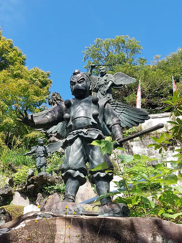 Statue impressionnante d'un guerrier ailé, entourée de verdure, sous un ciel bleu éclatant.