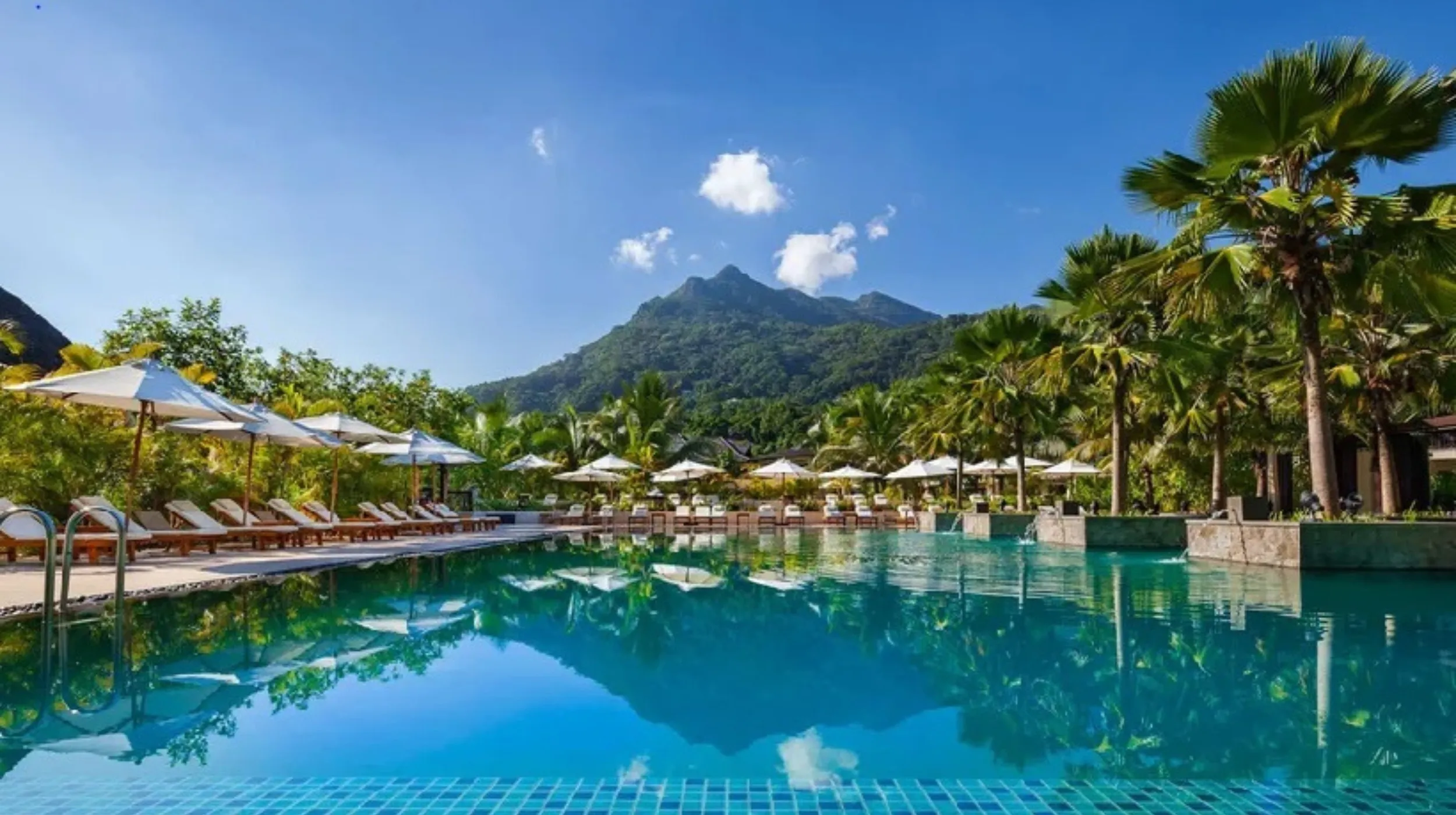 Piscine turquoise entourée de palmiers, avec une vue spectaculaire sur une montagne verdoyante.