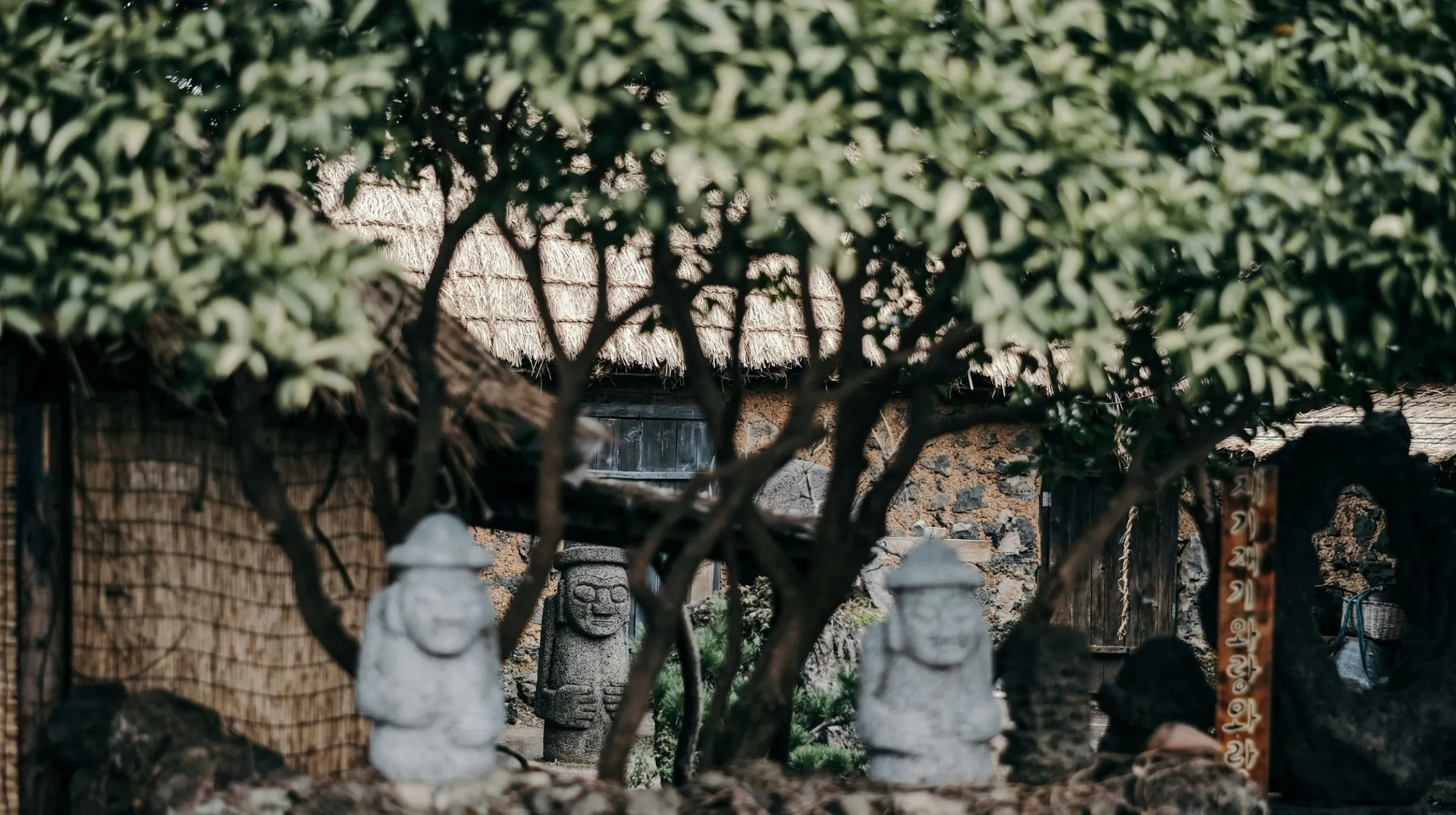 Scène paisible à Jeju en Corée du Sud, avec des statues de pierre entourées de verdure et d'une maison traditionnelle en arrière-plan.