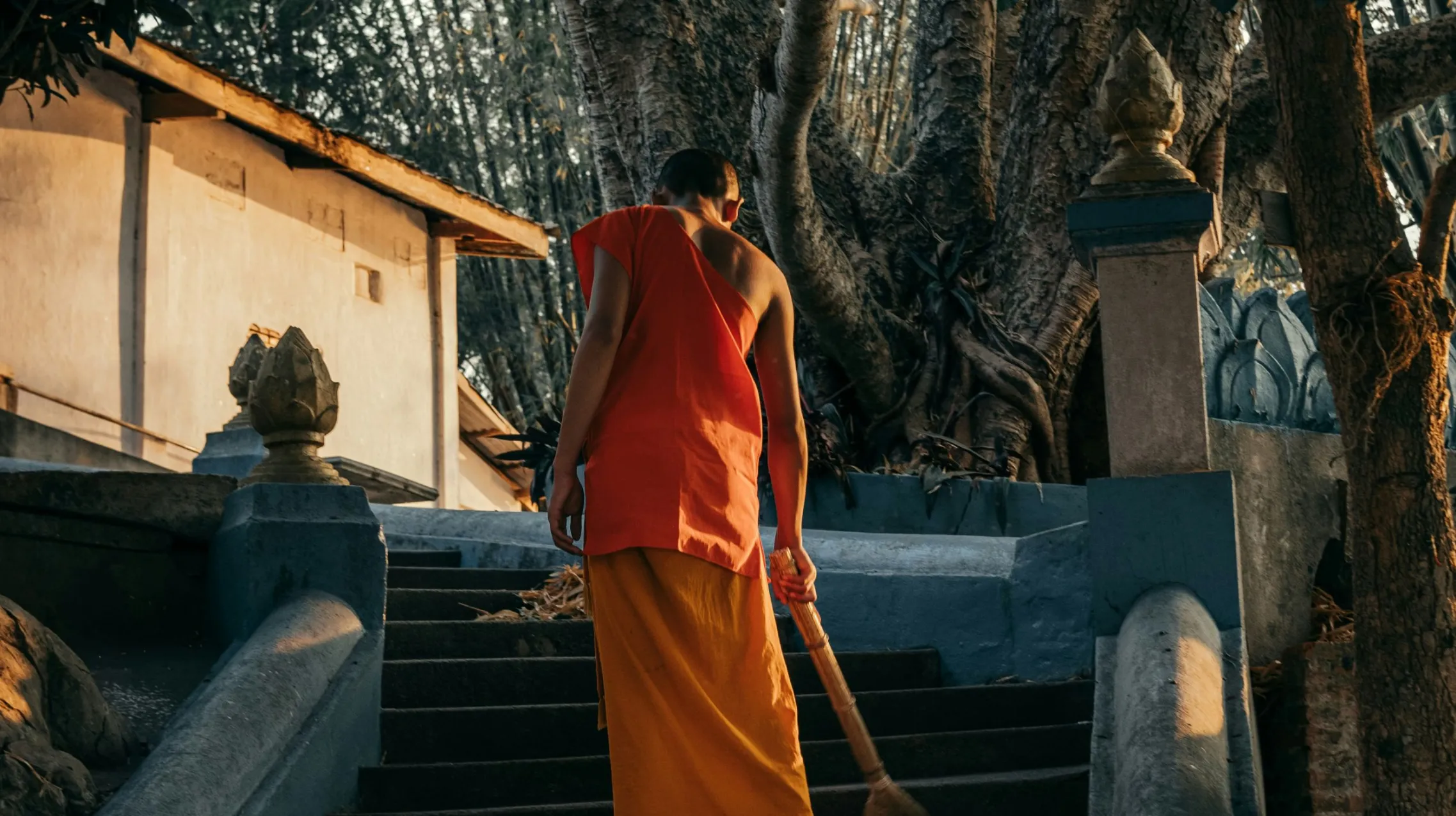 Moine en robe orange montant des escaliers, entouré d'une nature luxuriante et d'architecture traditionnelle.