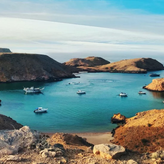 Côte paisible avec des bateaux au mouillage à Oman, entourée de collines arides et d'une mer turquoise.