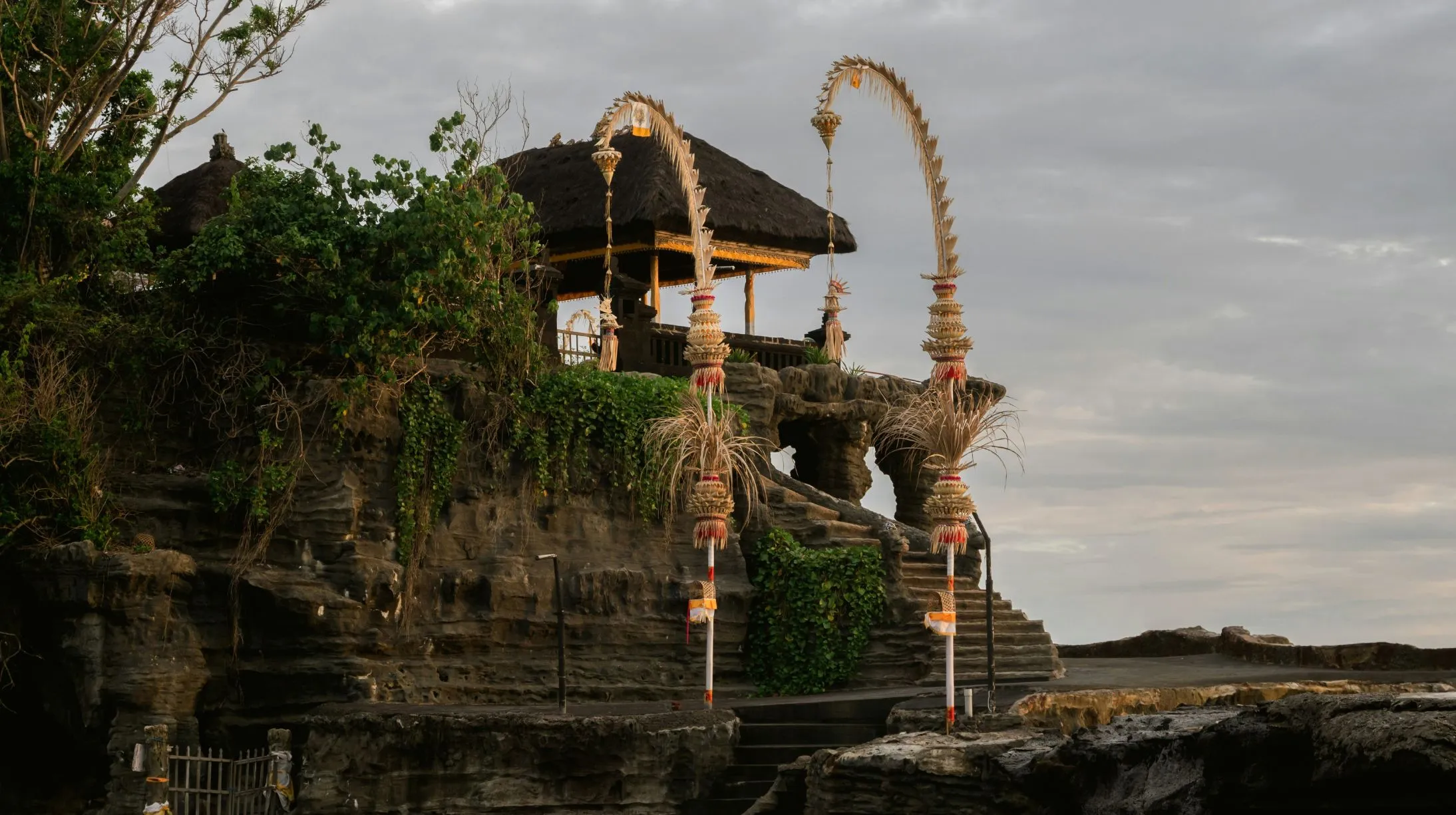 Temple de Tanha Lot enchanteur sur une falaise, entouré de verdure et de structures traditionnelles, sous un ciel nuageux.
