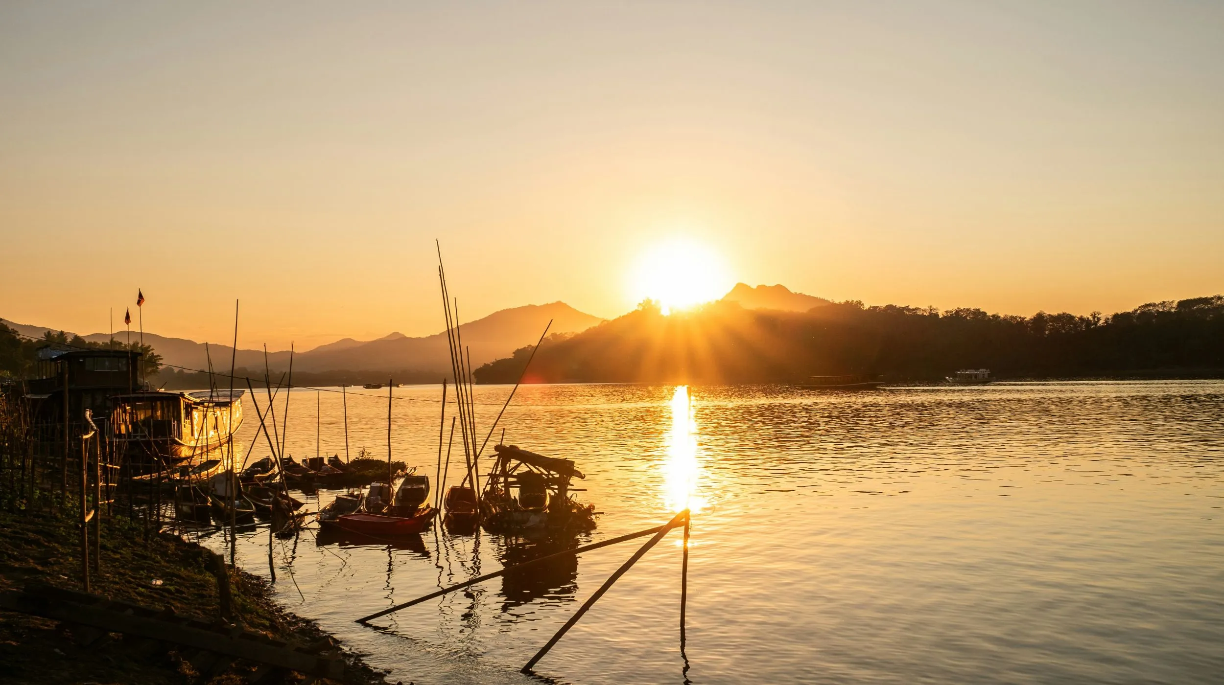 Coucher de soleil sur un fleuve, cadre paisible avec des bateaux colorés et des montagnes en arrière-plan.