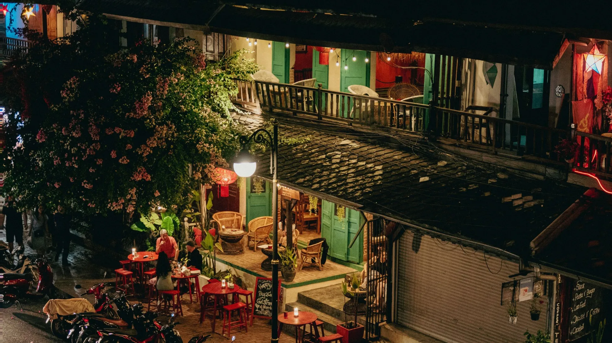 Scène animée d'une ruelle colorée avec des tables en bois, entourée de plantes luxuriantes et éclairée par des lanternes.